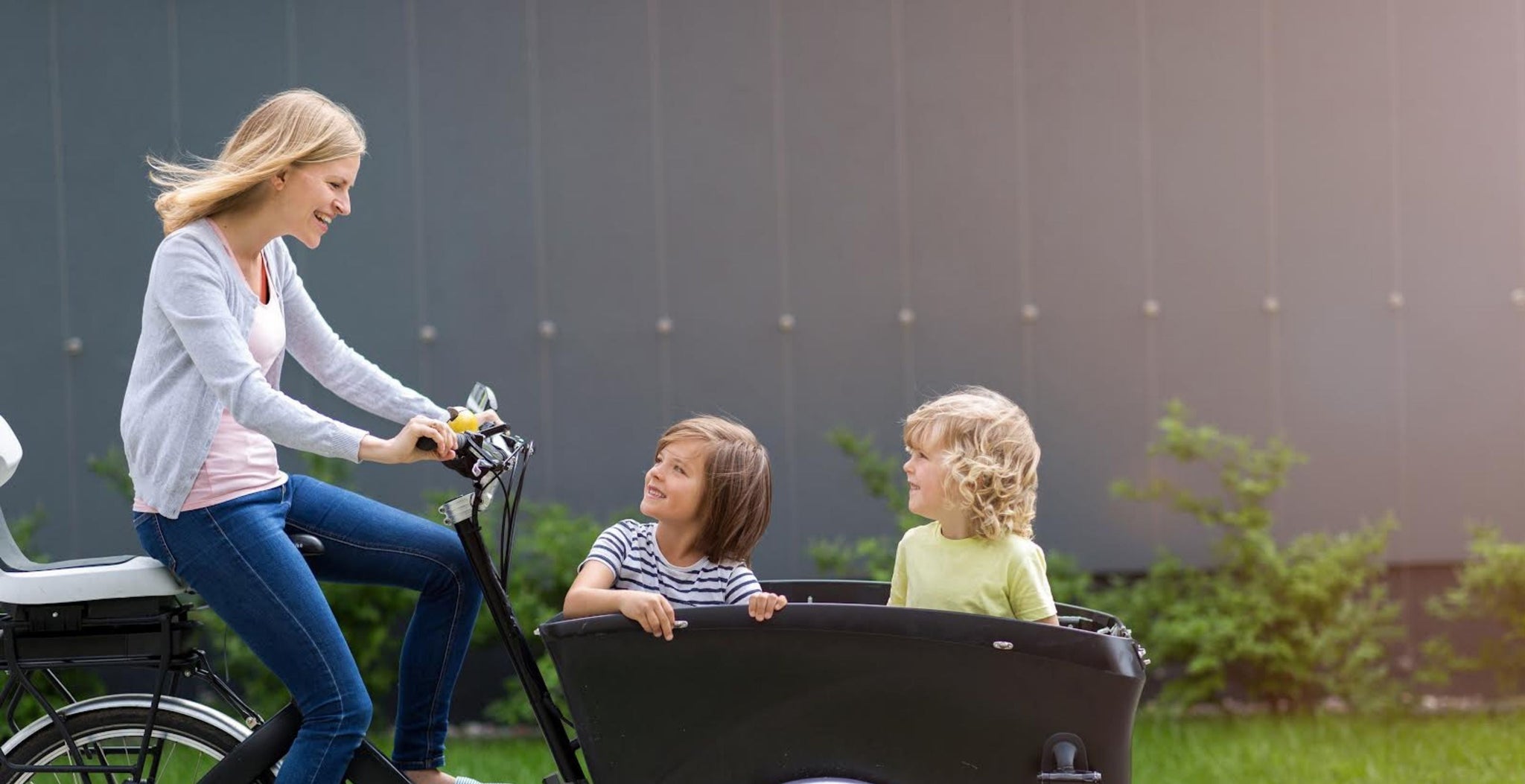 Woman riding a bicycle with two children in a cargo bike outdoors.