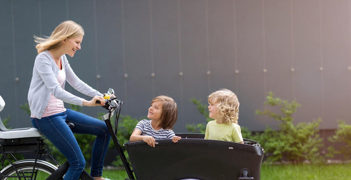 Woman riding a bicycle with two children in a cargo bike outdoors.
