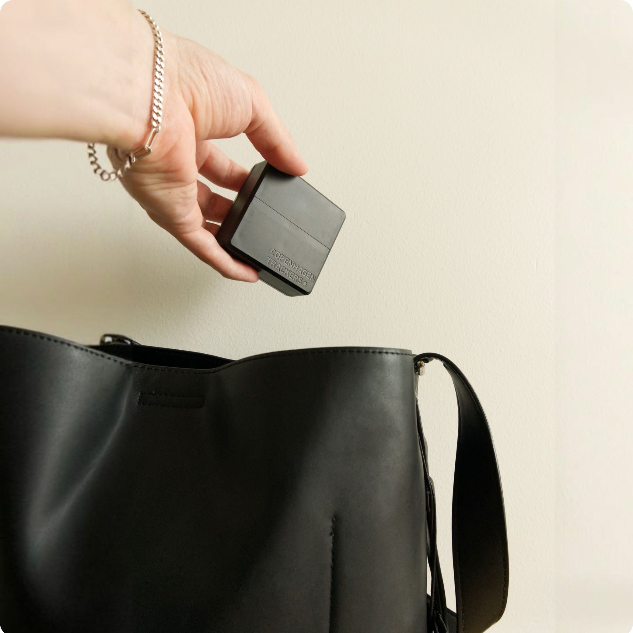 Hand holding the Cobblestone 2.0 GPS tracker above a black bag on a beige background