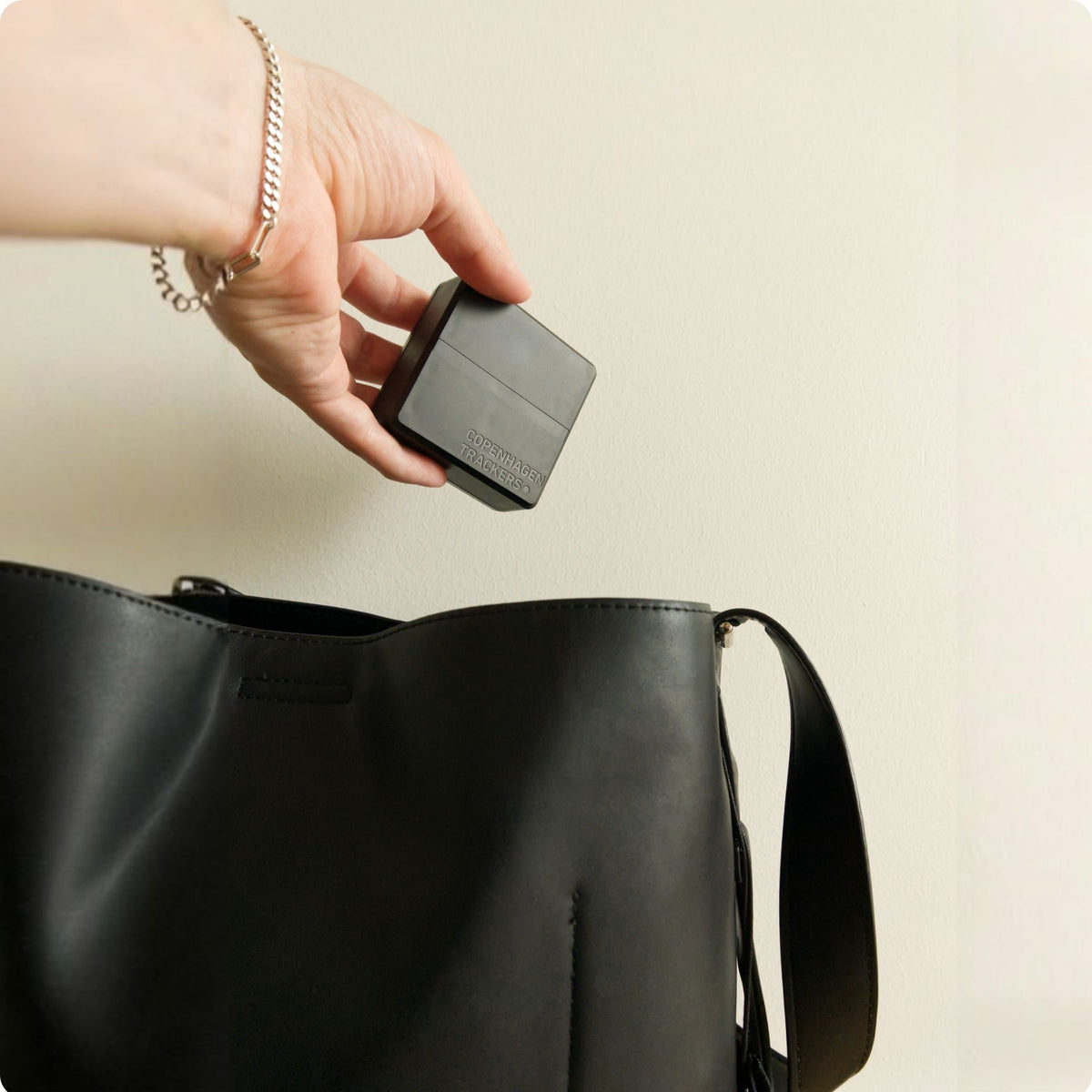Hand holding the Cobblestone 2.0 GPS tracker above a black bag on a beige background